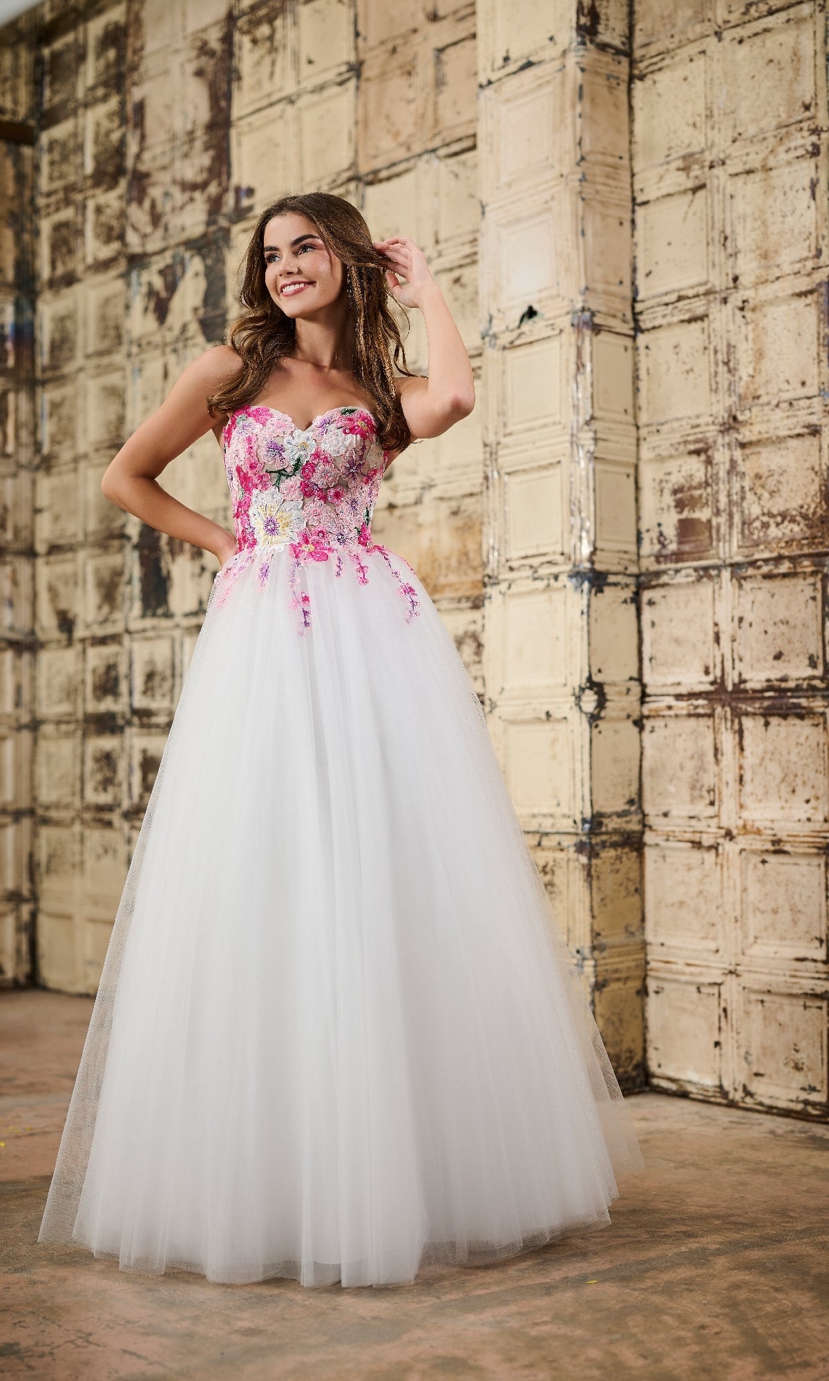 A woman smiles and touches her hair while posing in front of a textured wall, wearing the strapless Tiffany 16142 long prom dress with pink floral embroidery and a tulle skirt.